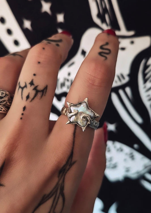 A close-up of a hand wearing a silver ring with a large, organic, star-like or mushroom-shaped setting. The hand has black tattoos and red nail polish, with a black and white graphic background.