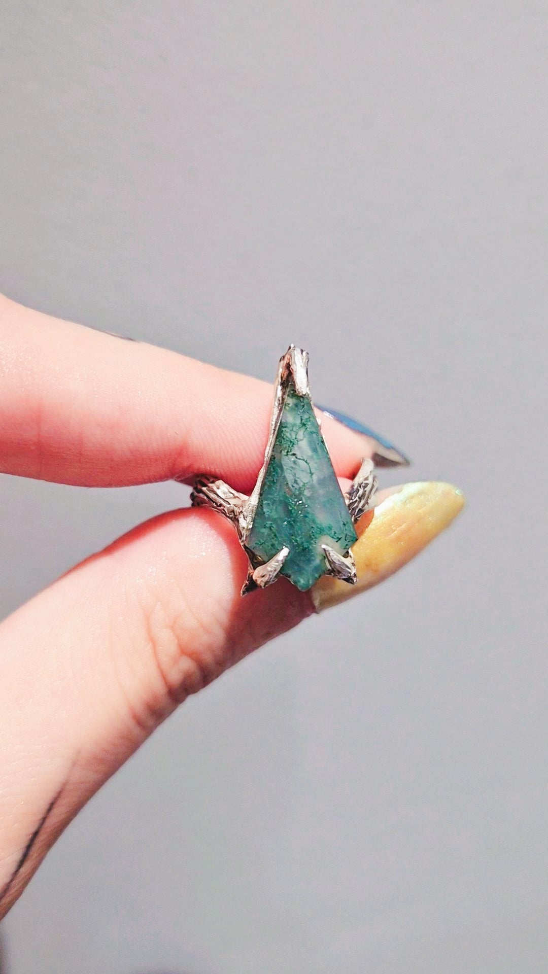 A hand holding a silver ring featuring a large, faceted green gemstone with a textured, moss-like surface.