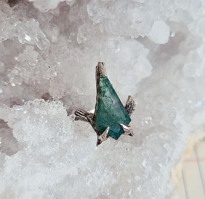 A silver ring featuring a triangular green stone inlay, displayed on a bed of white and pale pink crystal clusters.