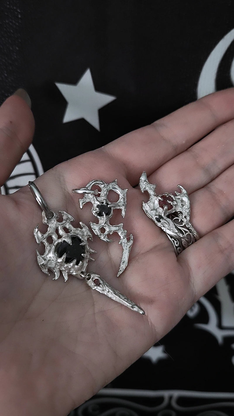 A hand holding three gothic silver-toned metal charms, including a ring with a skull and dagger motif, against a black background with white graphic elements. Cybersigilism earrings, silver cybersigilism earrings, silver cybersigilism jewelry, silver cybersigilism jewellery