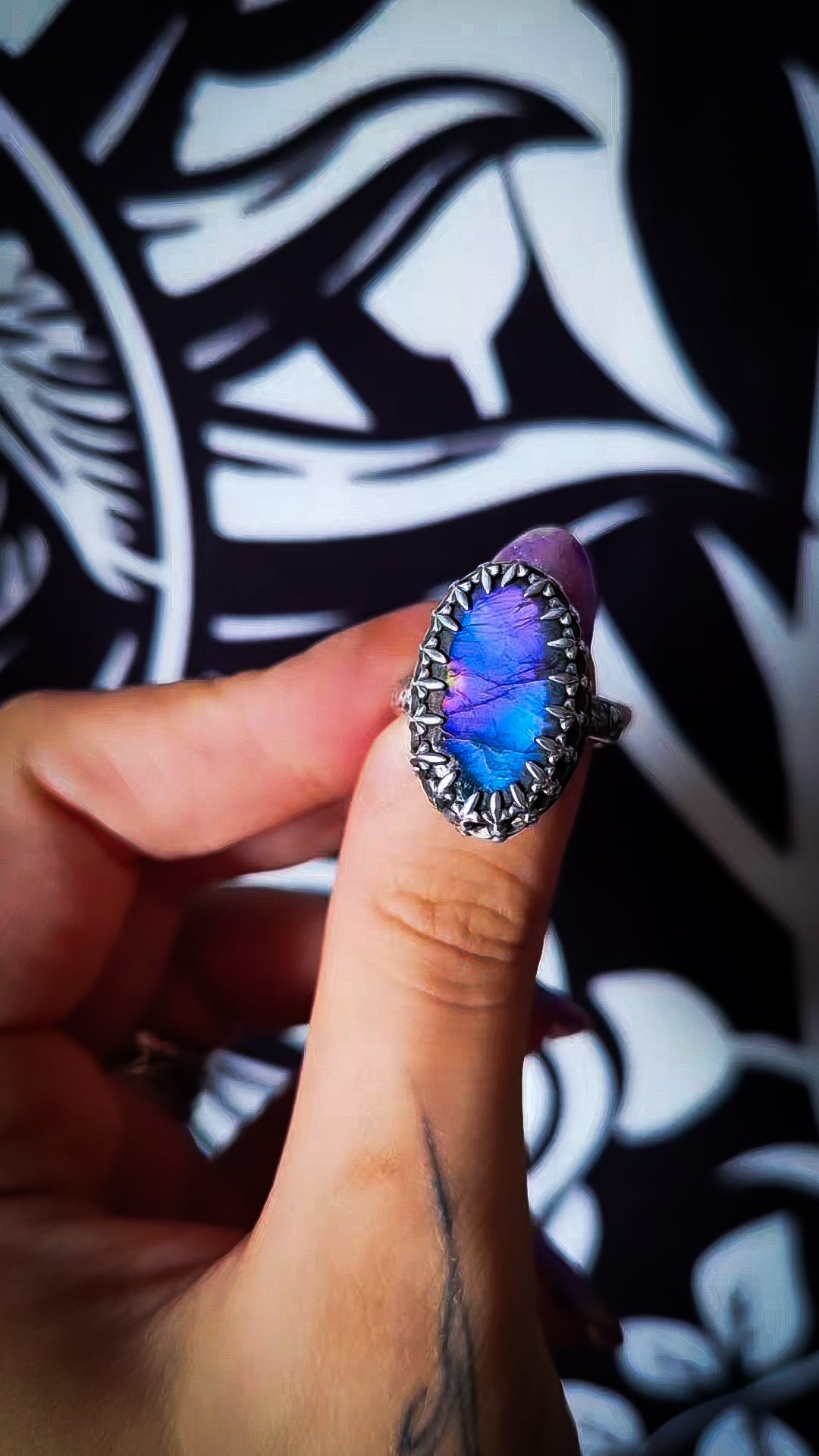 A close-up of a hand holding a silver ring with an oval iridescent blue and purple gemstone, set against a black and white patterned background.