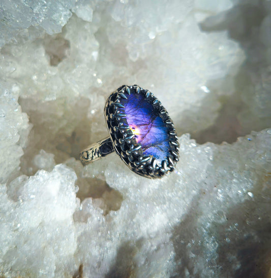 A silver ring with a large oval labradorite stone displaying a vibrant purple and blue play-of-color, resting on a bed of white crystal.