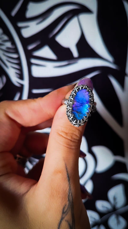 A close-up of a hand wearing a silver ring with a vibrant blue iridescent opal-like center stone, set against a black and white patterned background.