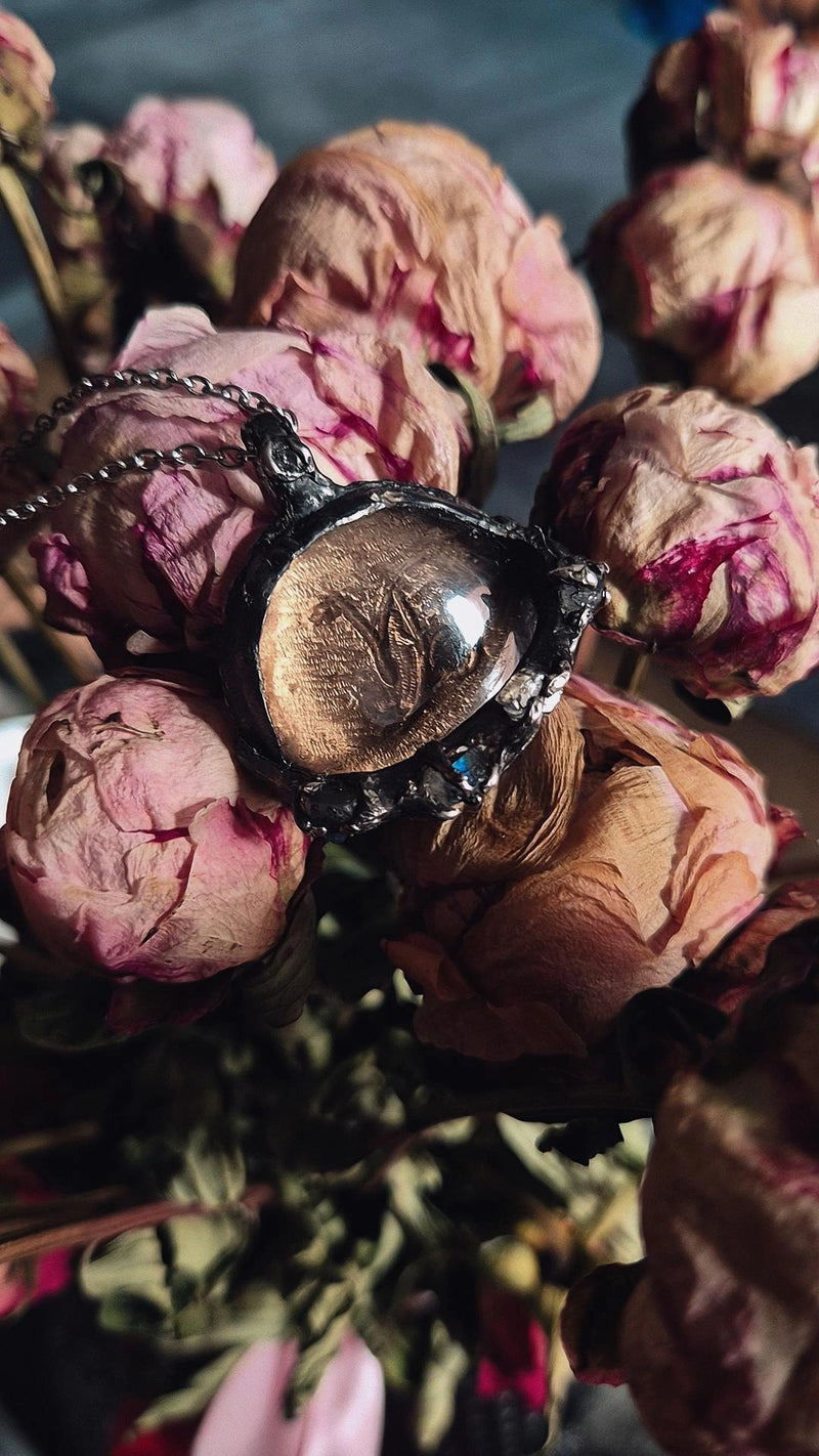 A close-up of a dark gothic-style pendant necklace with a chain, featuring a smoky quartz cab with moonstones resting on dried pink roses. Capricorn pendant, silver zodiac pendant