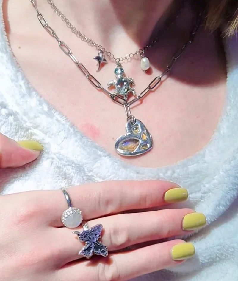 Close-up of a person wearing a silver necklace with a heart pendant and a butterfly ring on a white background.