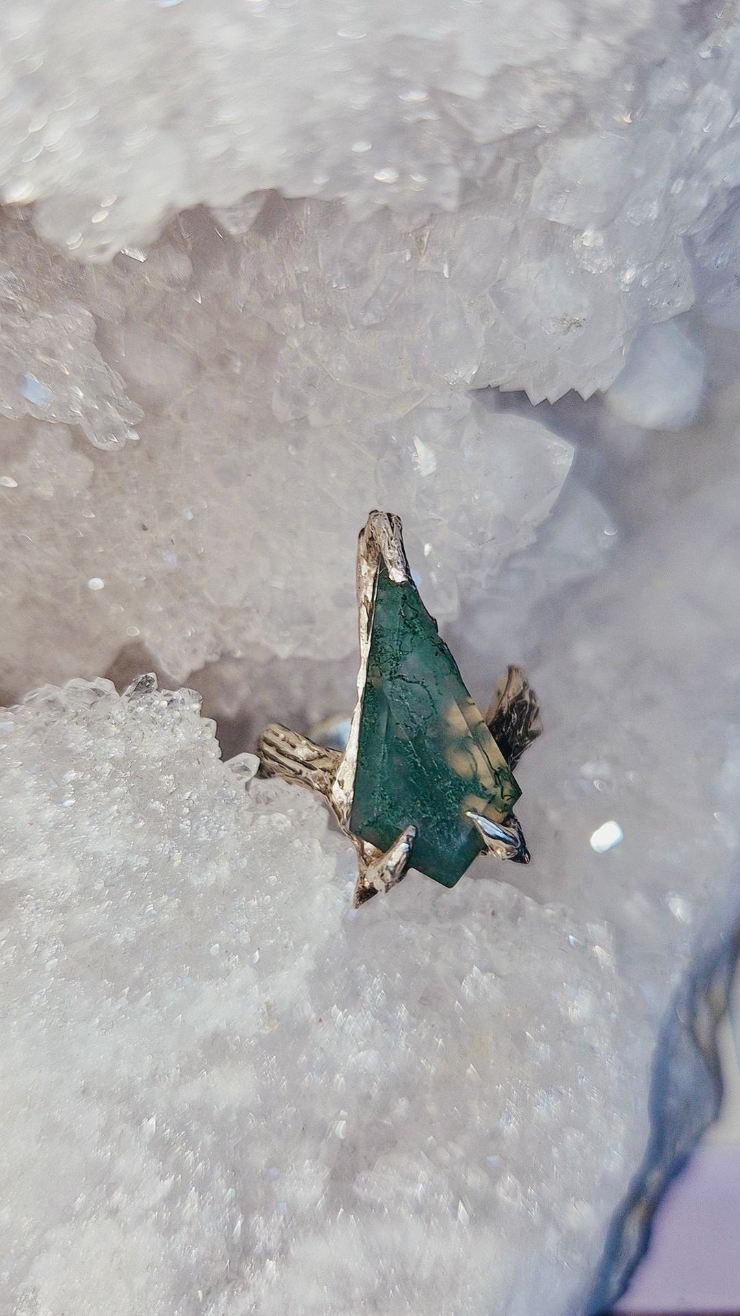 A coffin moss agatw gemstone ring with gold-toned metal settings, resting on a bed of white quartz crystal geode formations.