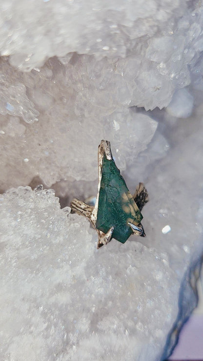 A coffin moss agatw gemstone ring with gold-toned metal settings, resting on a bed of white quartz crystal geode formations.
