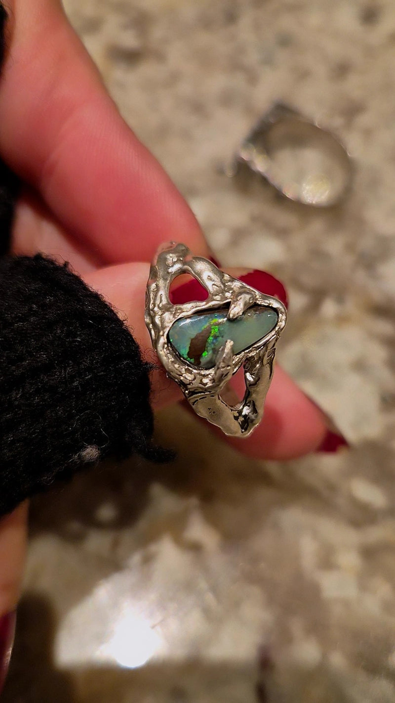 Silver ring with a heart-shaped green gemstone held by a hand on a textured surface.