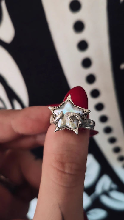 A silver-toned ring with a star-shaped setting and a white inlay, shown being worn on a finger with red nail polish against a black and white patterned background.