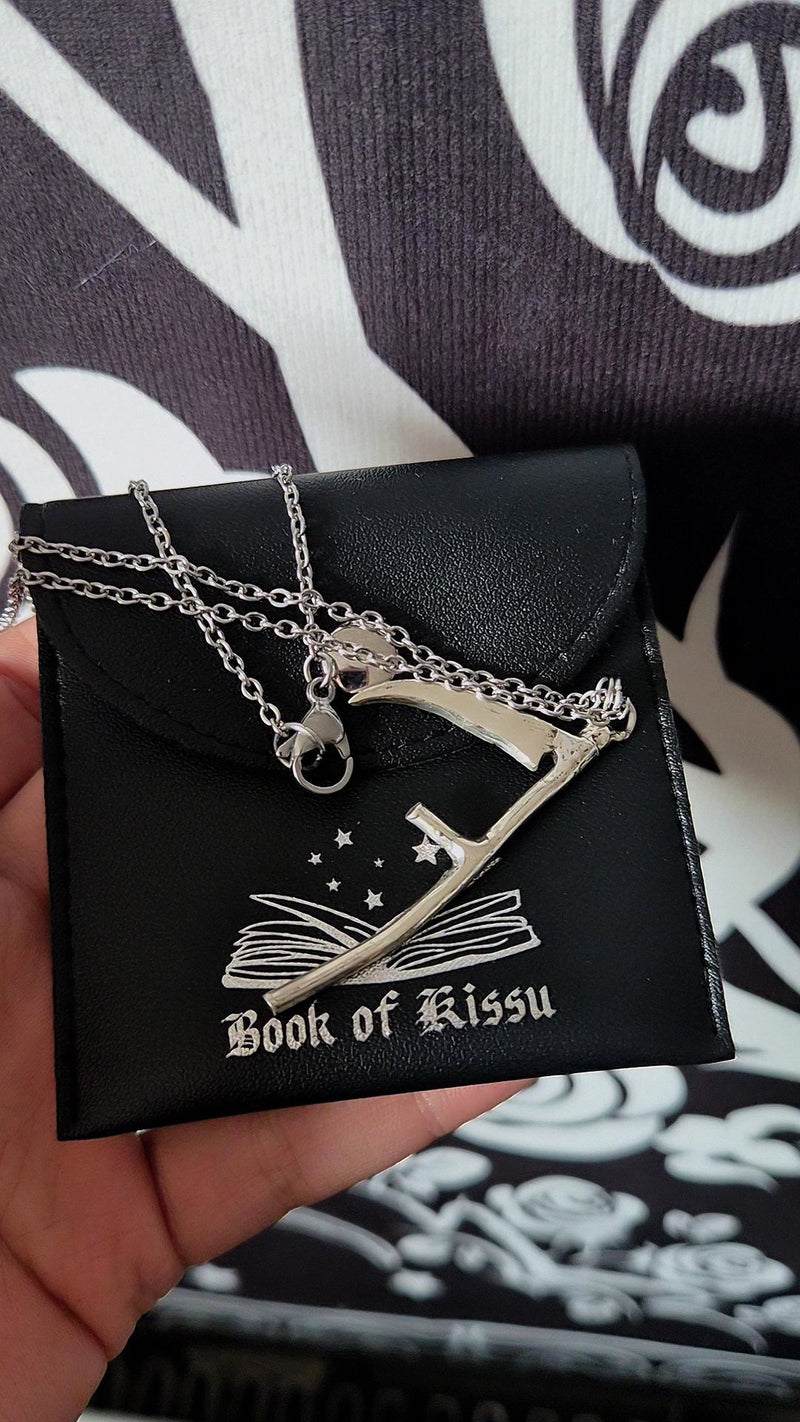 Silver scythe pendant on a black card labeled 'Book of Kissu' against a black and white patterned background.