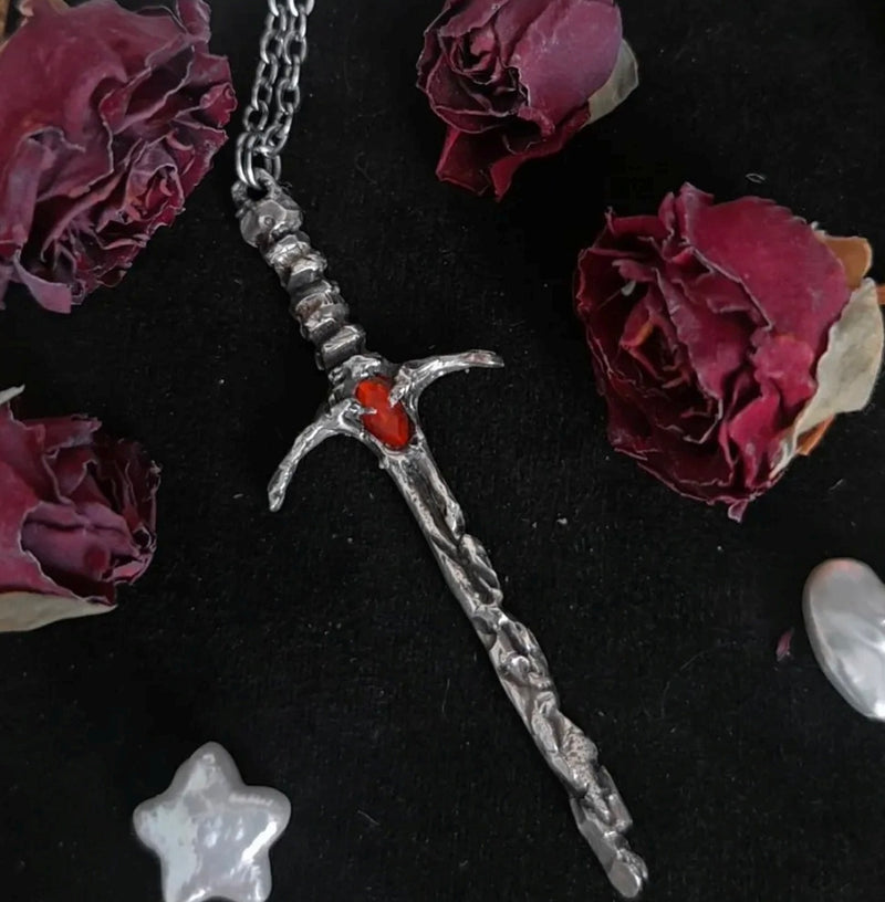 Silver sword-shaped pendant with a garnet gemstone on a dark surface with dried roses.