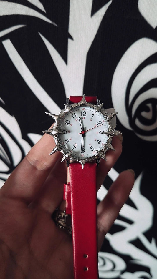 Watch with a red strap and spiky design held by a hand against a black and white patterned background.