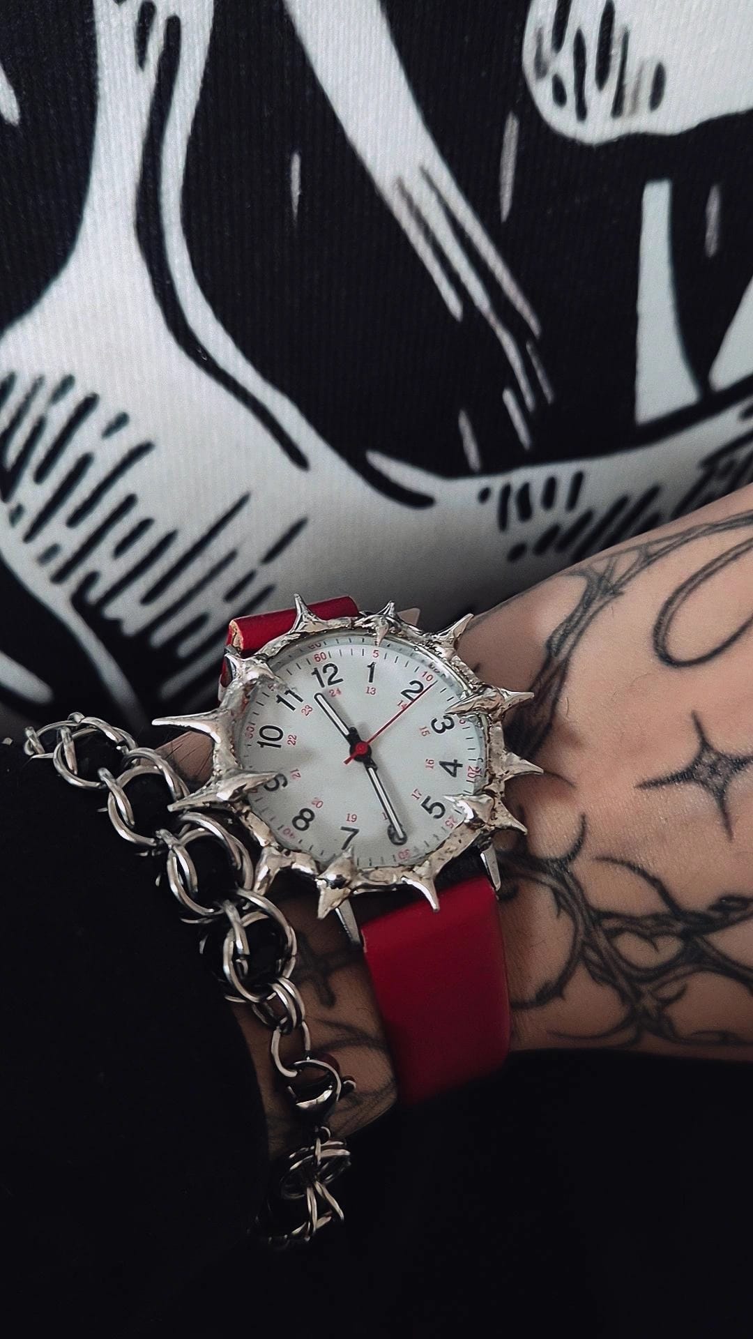 Spiked wristwatch with red strap worn by a person with tattoos on a black and white patterned background.