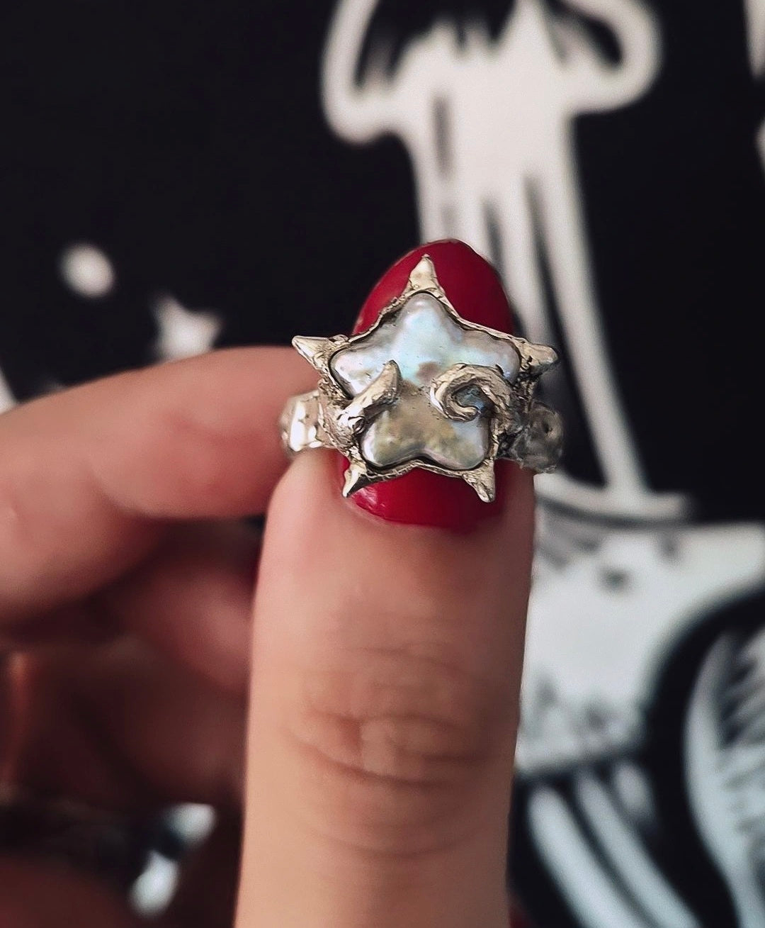 A close-up of a hand with red nail polish holding a silver-toned ring featuring a white mother-of-pearl star-shaped inlay.