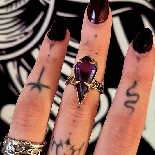 Close-up of a hand with dark nail polish and a bronze amethyst ring against a black and white patterned background.